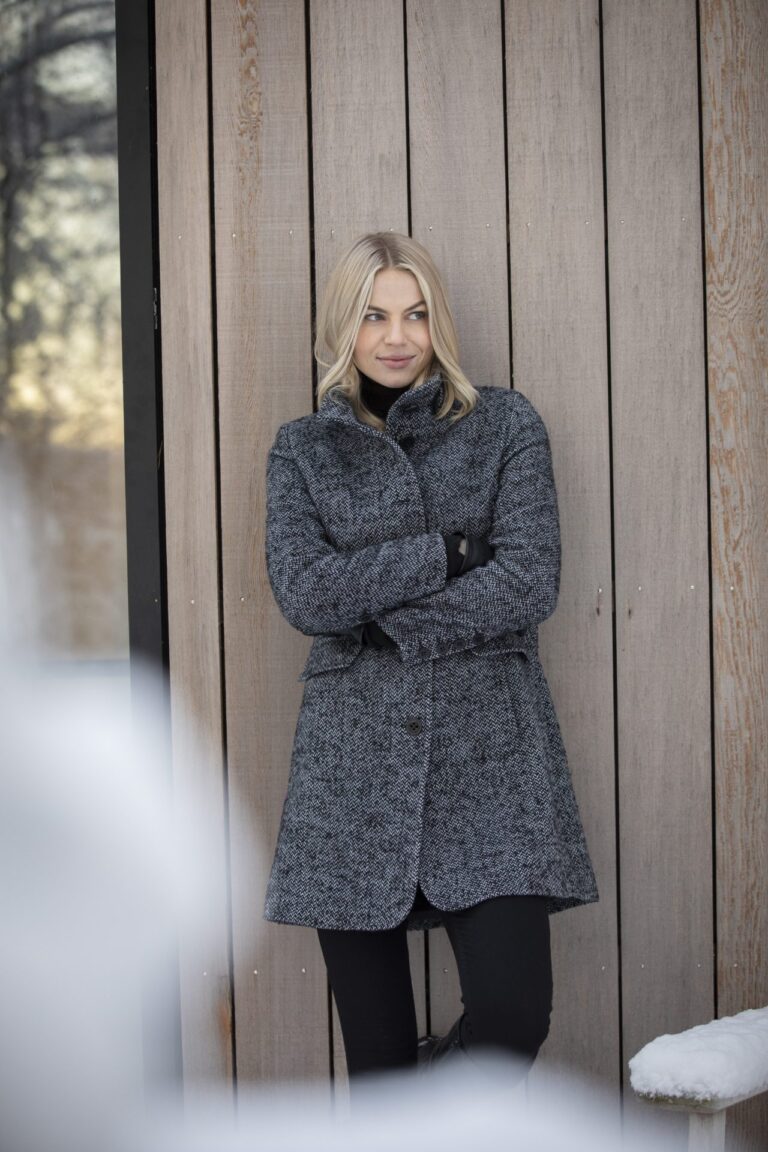 L'image montre une femme au cheveux blonds ayant un beau veston et un pantalon noir. De plus, ceci se passe durant l'hiver et l'arrière plan c'est du bois d'une maison.