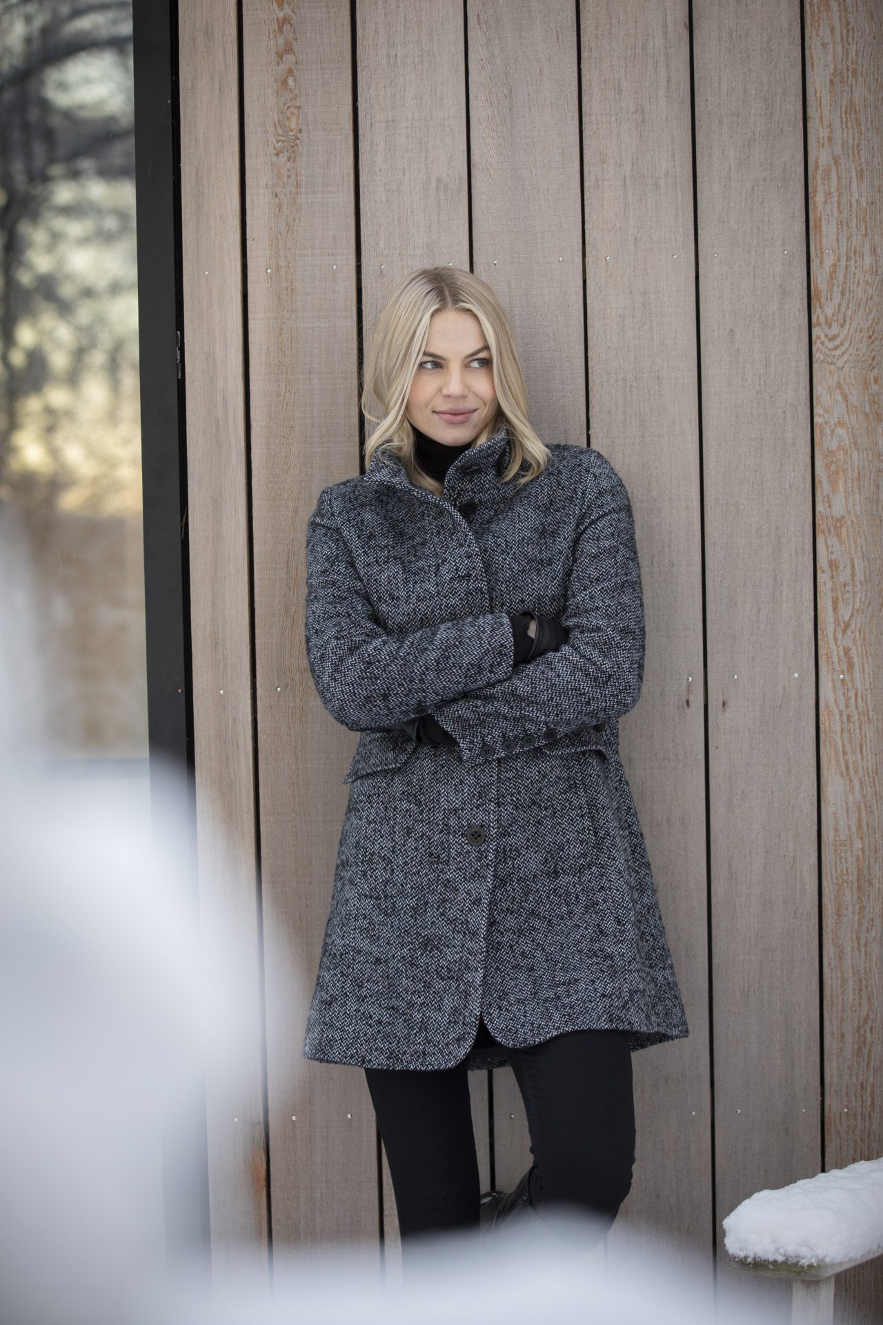 L'image montre une femme au cheveux blonds ayant un beau veston et un pantalon noir. De plus, ceci se passe durant l'hiver et l'arrière plan c'est du bois d'une maison.