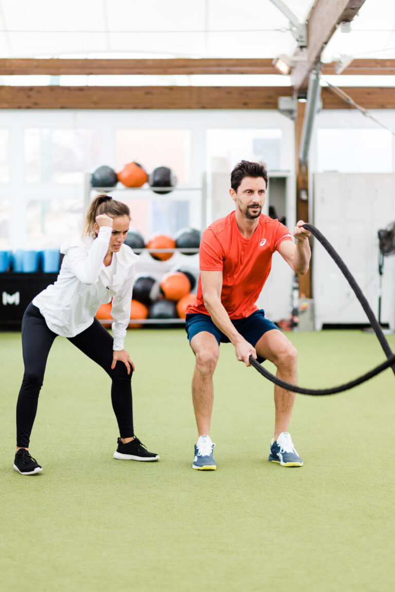 L'image montre une entraîneuse qui entraîne un homme à devenir plus fort dans une salle de musculation. L'homme fait de la corde.