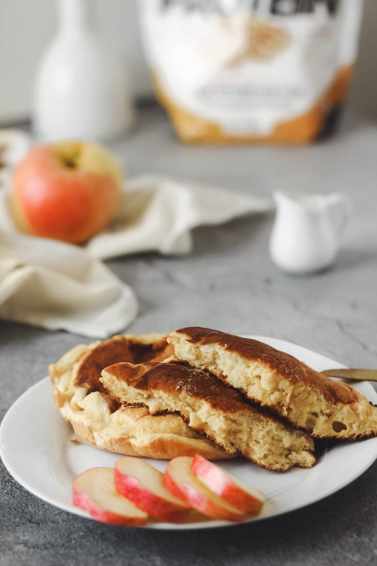 L'image montre un déjeuner contenant des pancakes, des pommes et en arrière plan, il y a une pomme avec une petite tasse à sirop d'érable avec un sachet de farine.
