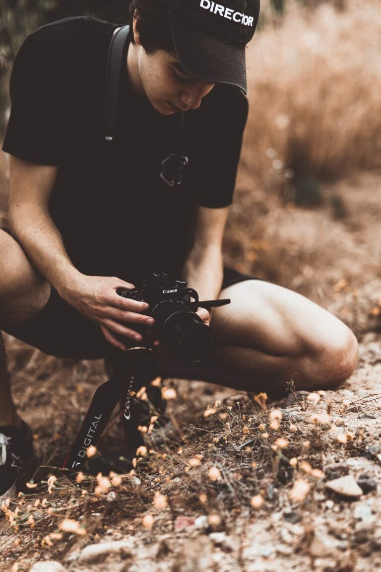 L'image montre un homme agenouiller sur de la terre contenant plusieurs branches mortes autour. L'homme porte une casquette noir et des vêtements noirs et celui-ci tient une caméra dans les mains.